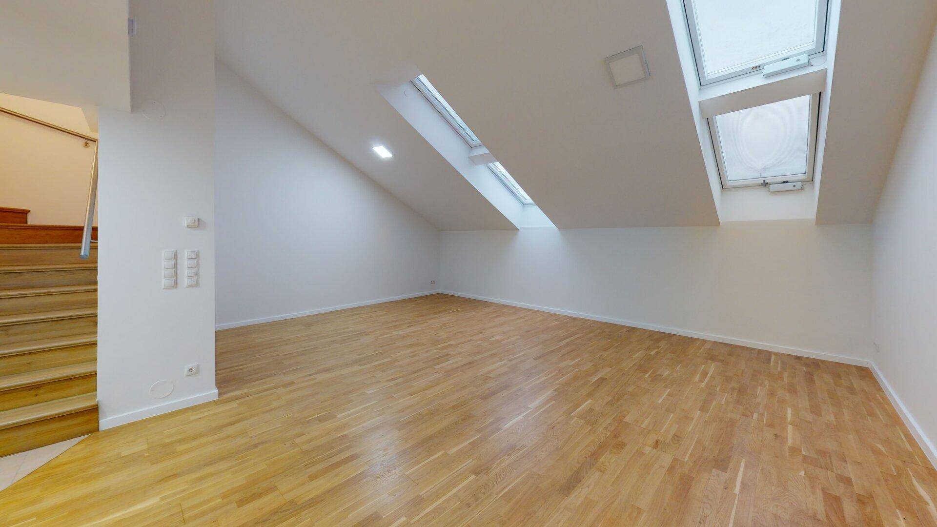 Heller Wohnraum mit Holzboden, Dachschrägen und mehreren Dachfenstern, angrenzend an eine Treppe.