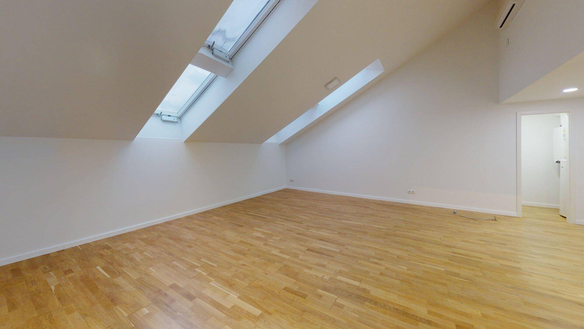 Geräumiger Raum im Dachgeschoss mit hellem Holzboden und schrägen Wänden, durch Dachfenster belichtet.