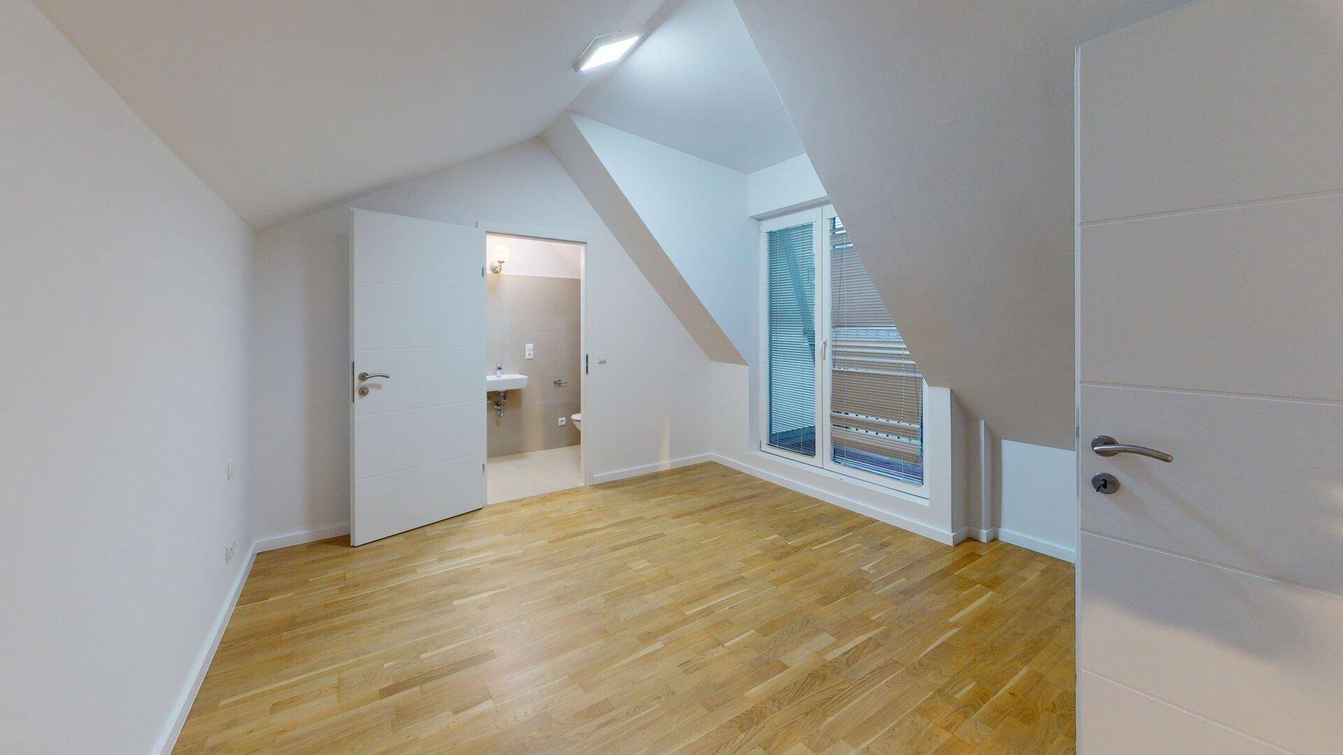 Schlafzimmer mit Holzboden, Dachschräge und Zugang zu einem Badezimmer sowie einem Balkon.