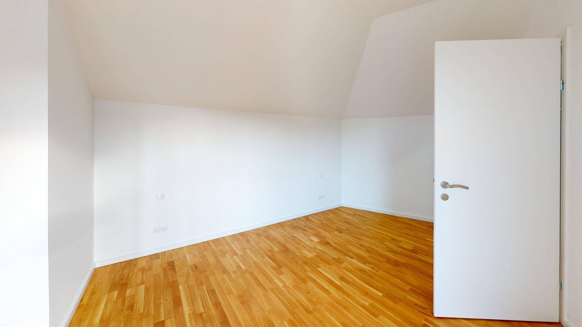 Leeres Zimmer mit Holzboden, weißen Wänden und schräger Decke, bereit zur individuellen Gestaltung.