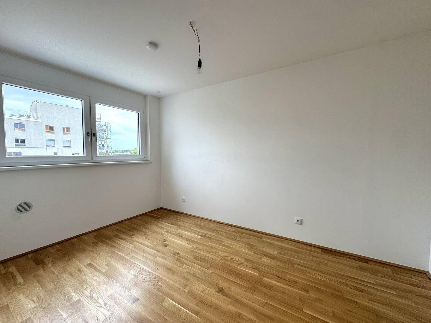 Helles, leeres Zimmer mit Holzboden und einem großen Fenster, das viel Tageslicht hereinlässt.
