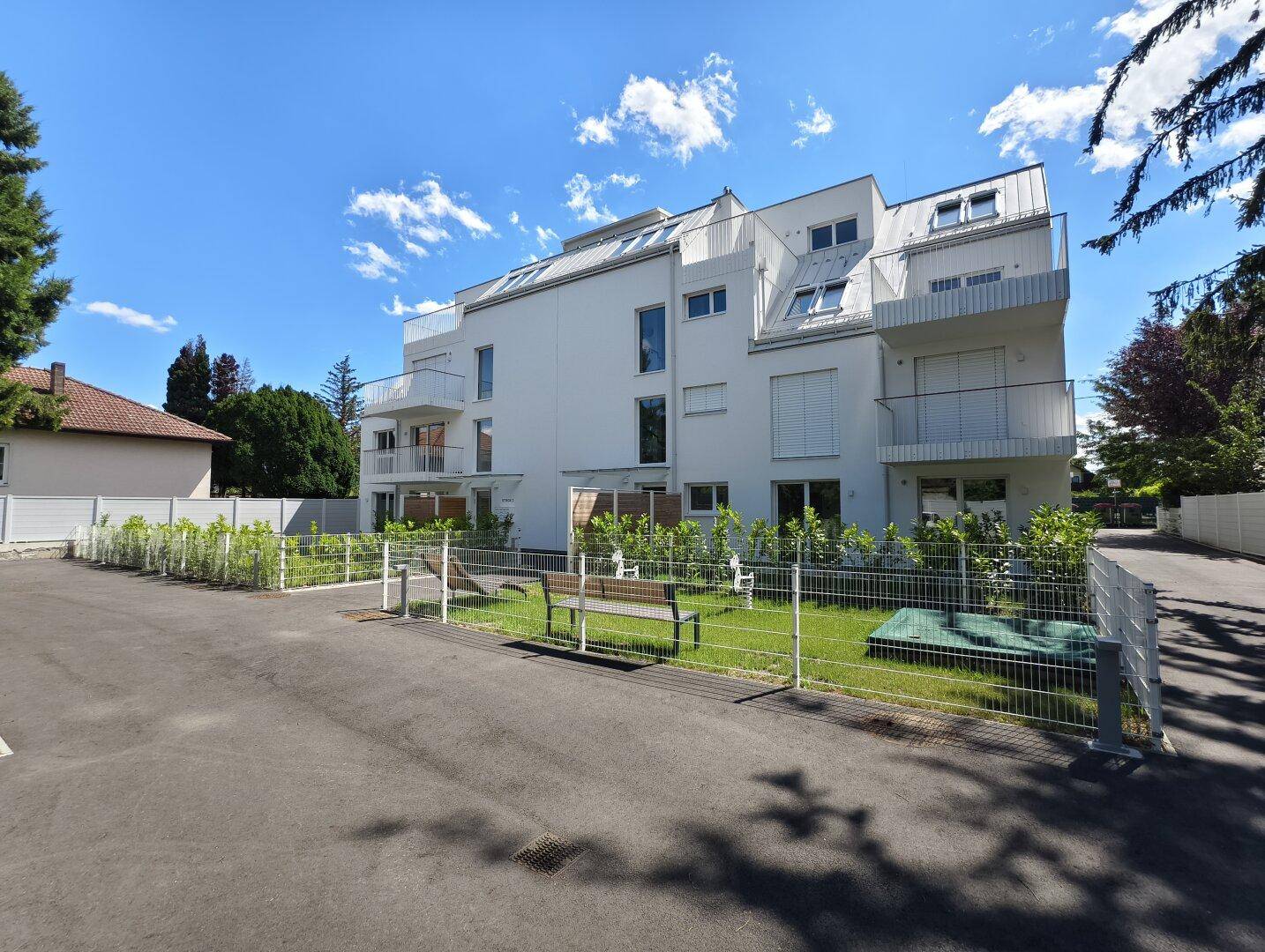 Ansicht der modernen Wohnanlage mit Gemeinschaftsgarten, Sitzbank und Parkmöglichkeiten im Freien.