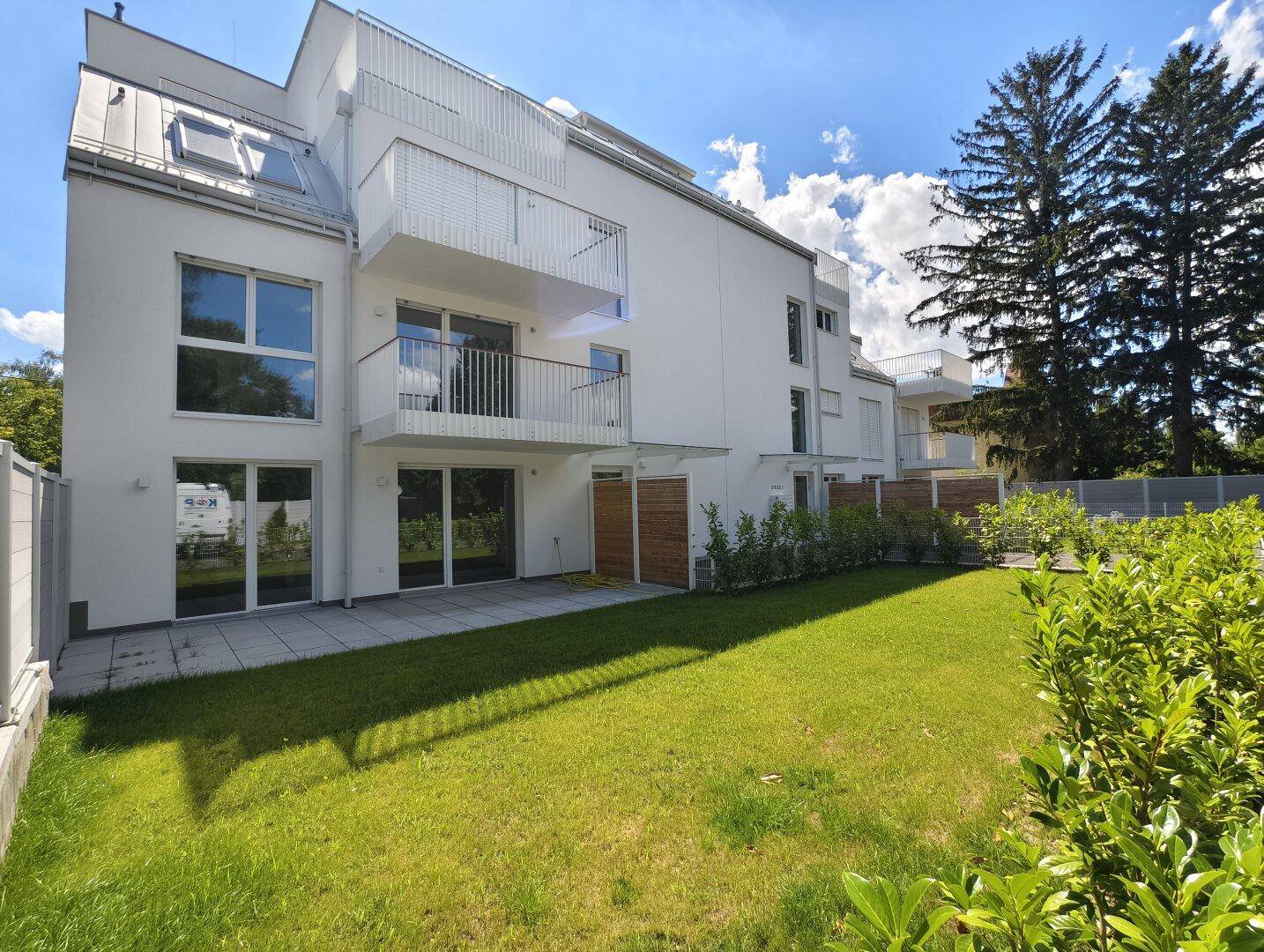 Rückseite des Gebäudes mit gepflegtem Rasen, Terrasse und mehreren Balkonen, umgeben von Grün.