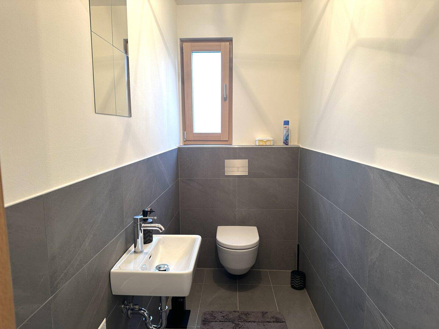 Modernes Gäste-WC mit grauen Fliesen, wandhängendem WC und kleinem Waschbecken.