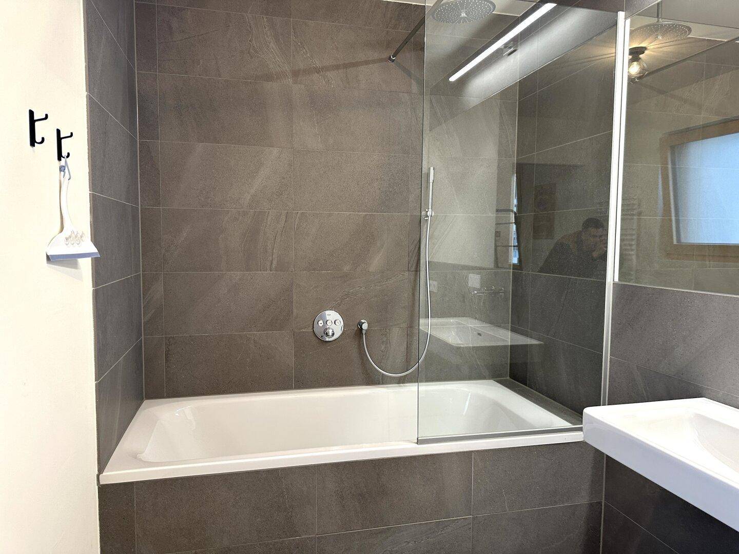 Zeitgemäßes Badezimmer mit grauen Fliesen, Badewanne und Duschbereich mit Glasabtrennung.