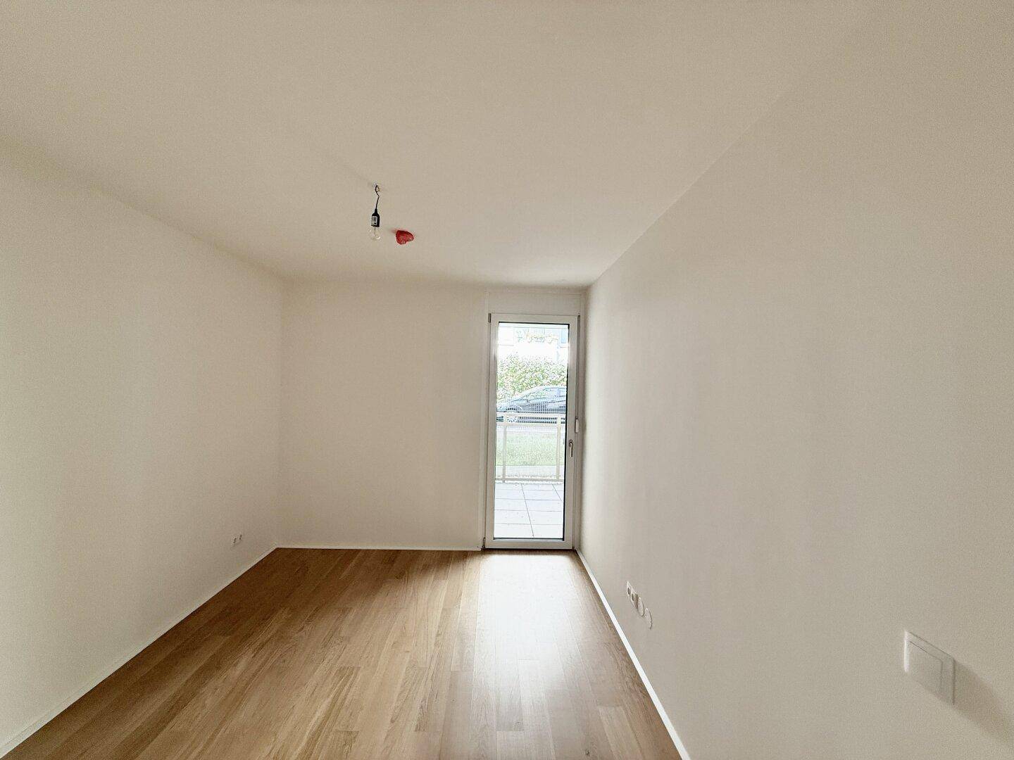 Leeres Zimmer mit Parkettboden und einer Tür, die zu einem Balkon oder Garten führt.