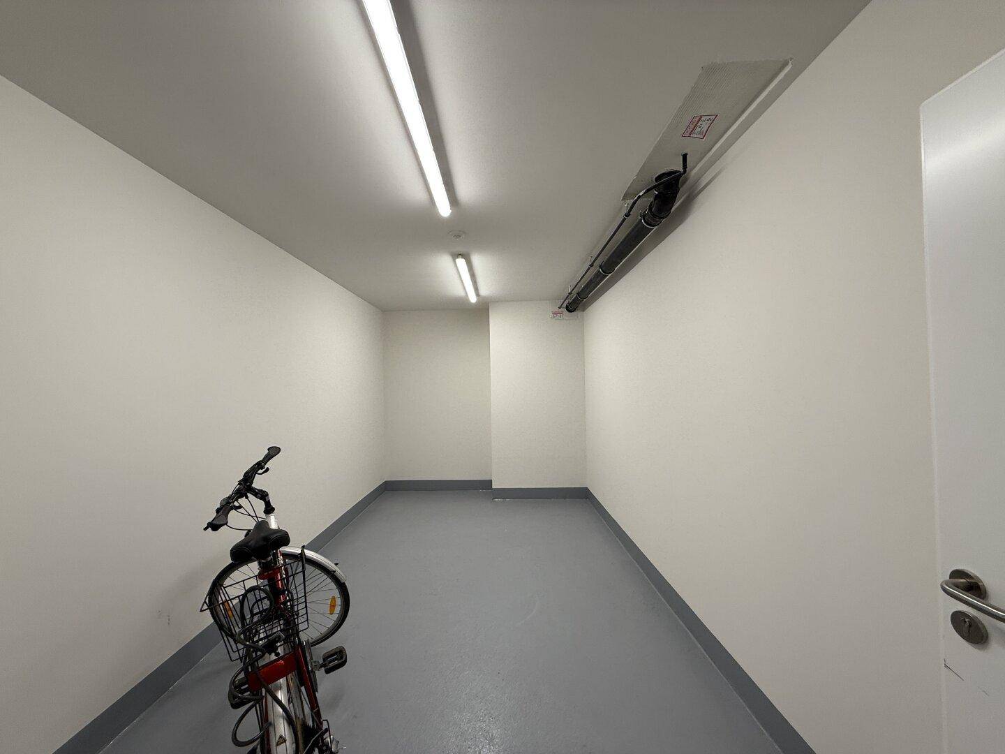 Heller Fahrradraum mit grauem Boden und weißen Wänden, ausgestattet mit Leuchtstoffröhren.