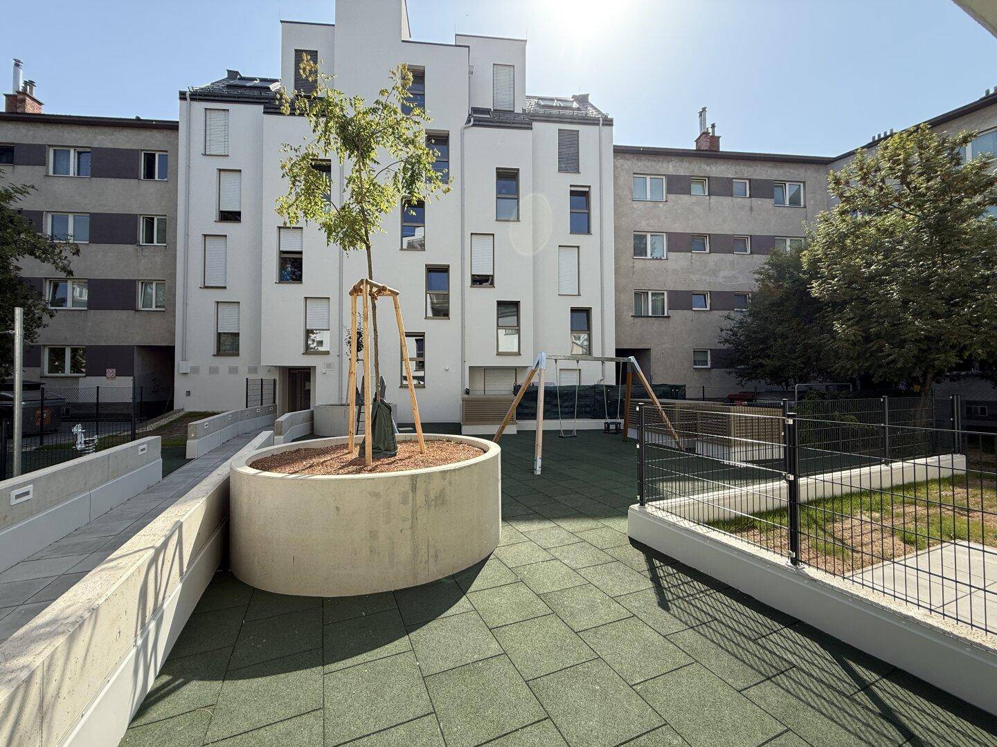 Moderner Spielplatz mit Schaukeln und einem Baum in einem Hochbeet, umgeben von Wohngebäuden.