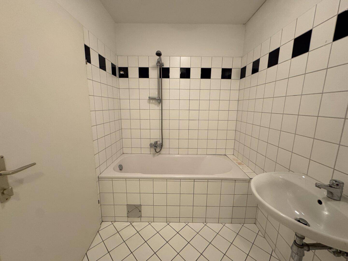 Badezimmer mit Badewanne, Waschbecken und weißen Fliesen mit schwarzem Akzentstreifen.