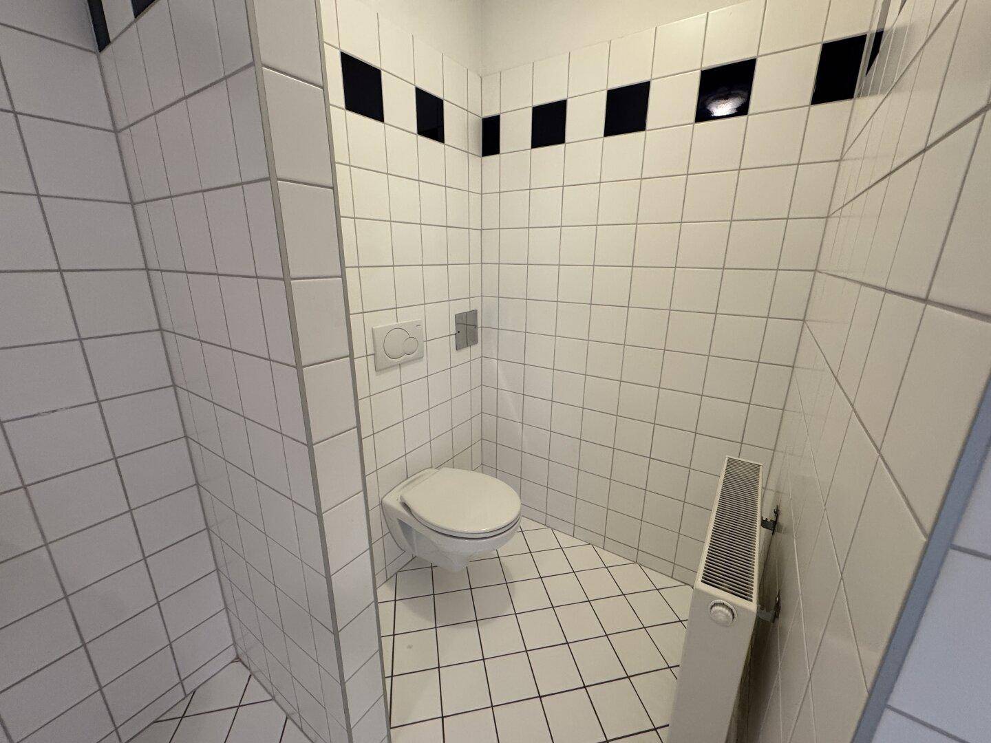 Separate Toilette mit Wand-WC, Heizkörper und weißen Fliesen mit schwarzem Akzent.