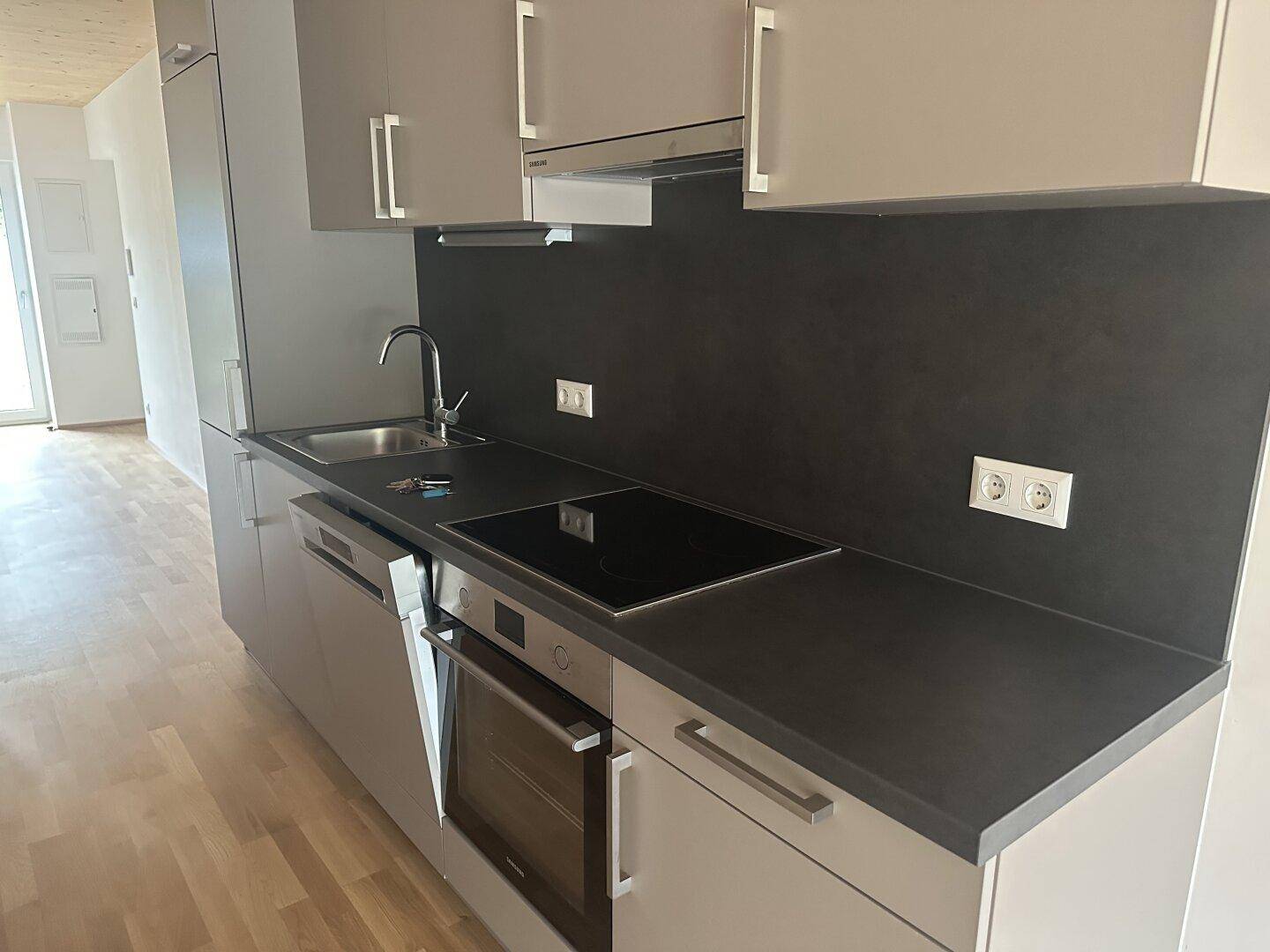 Moderne Einbauküche mit grauen Schränken, dunkler Arbeitsplatte und integrierten Elektrogeräten in hellem Raum.