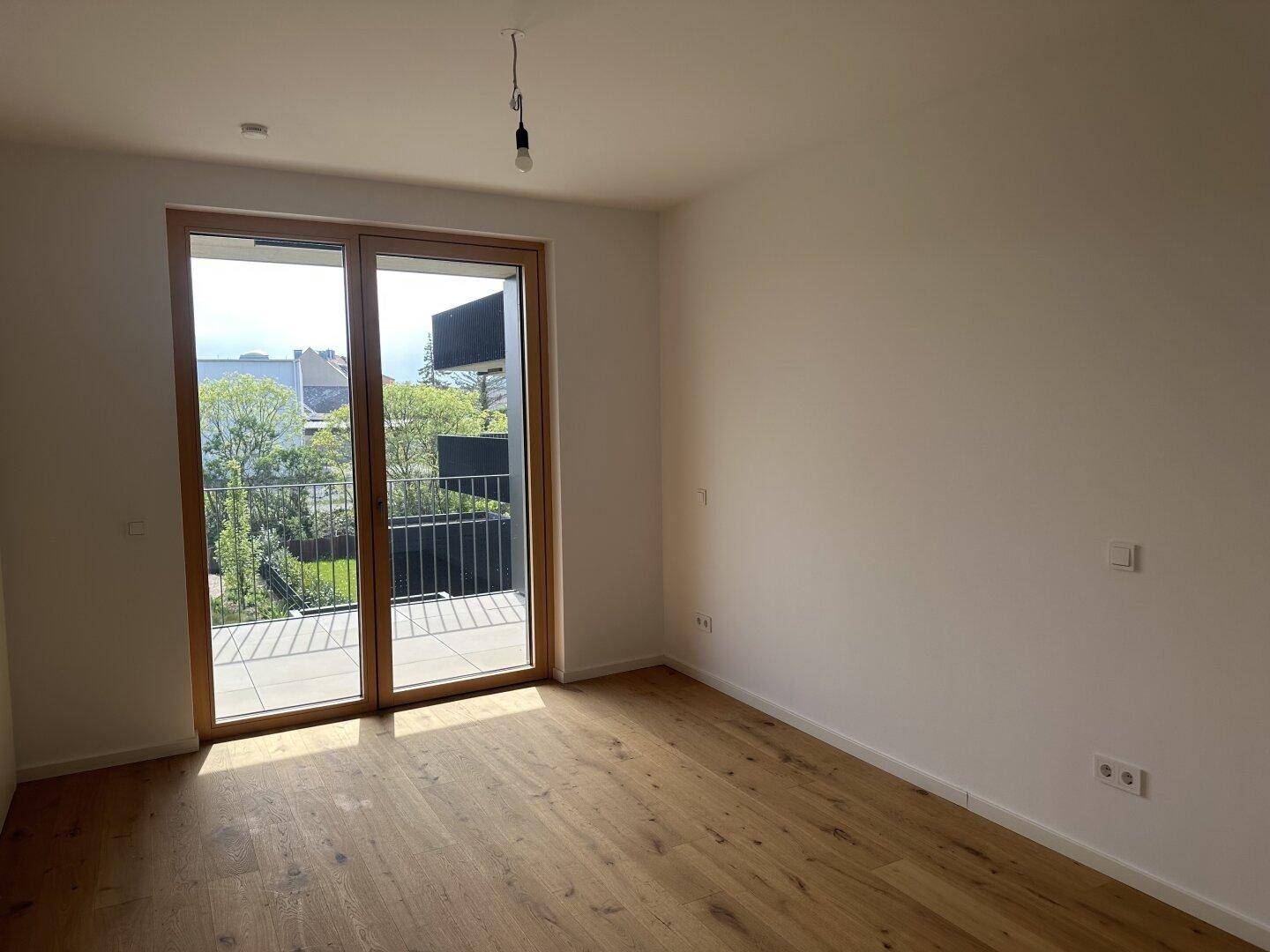 Heller Raum mit hochwertigem Holzboden und großem Balkonzugang, der einen grünen Ausblick bietet.