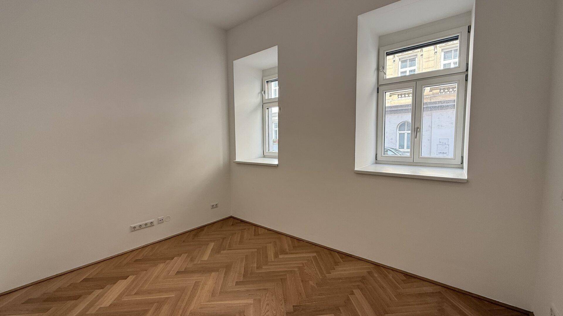 Leeres Schlafzimmer mit Fischgrätparkett, weißen Wänden und zwei Fenstern für natürliches Licht.
