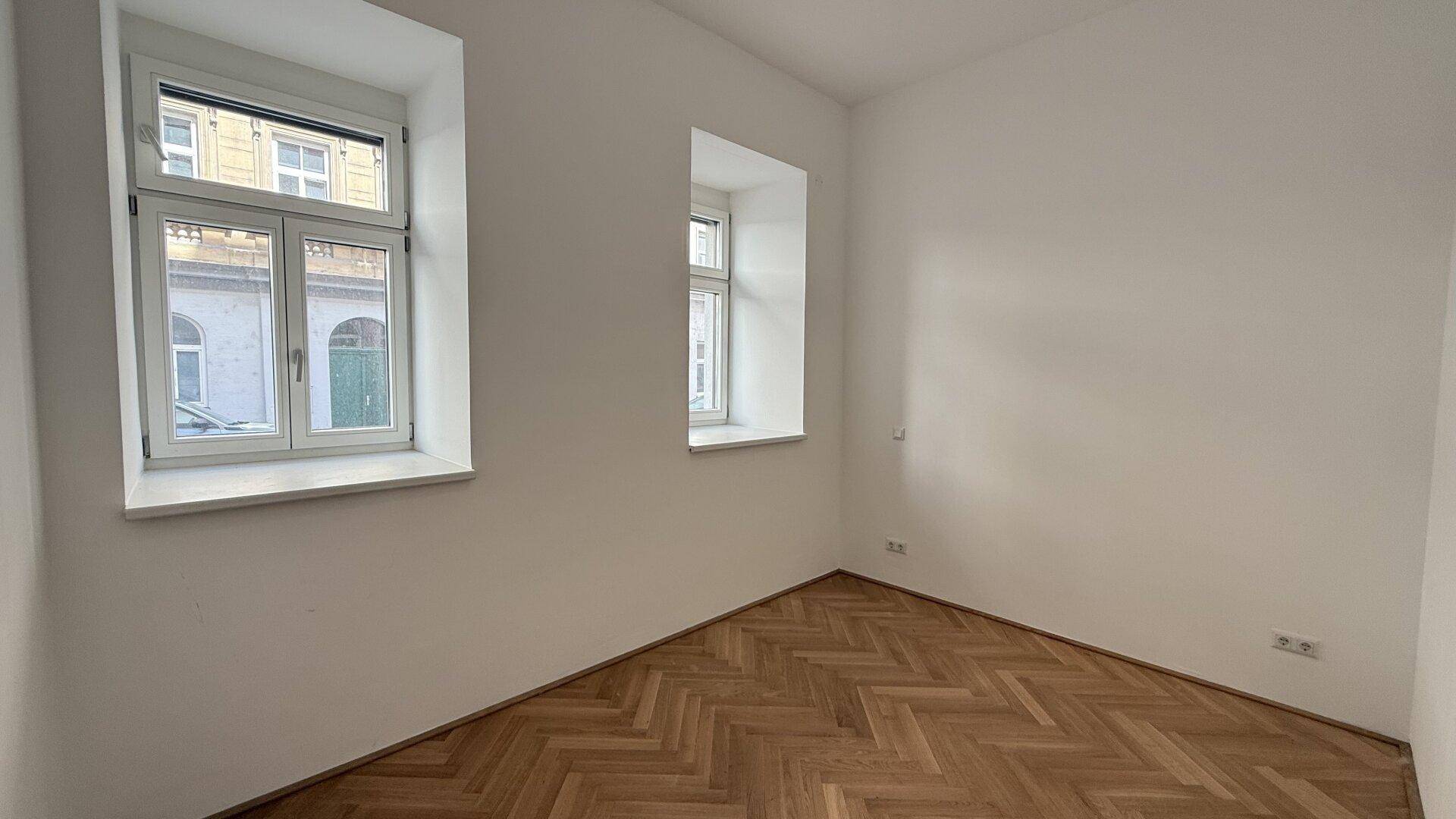 Geräumiges Zimmer mit Fischgrätparkett und zwei Fenstern, die einen Blick nach draußen bieten.