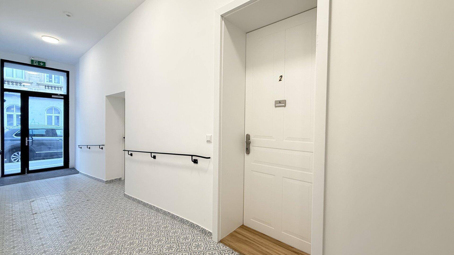 Heller Eingangsbereich des Gebäudes mit gemusterten Fliesen, Handlauf und Wohnungstür Nummer 2.