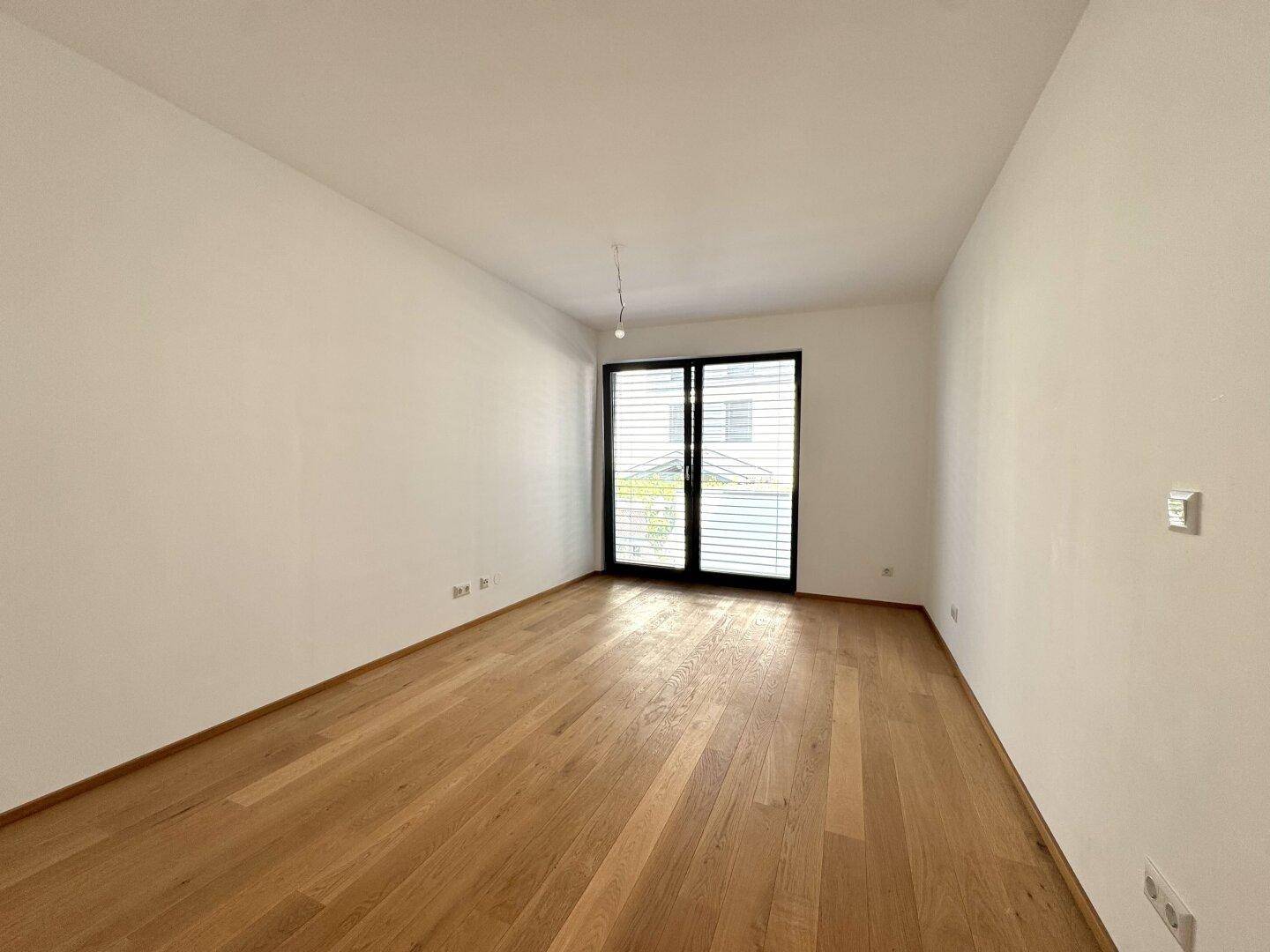 Helles Zimmer mit Holzboden und großem Fenster mit geschlossenen Rollläden, ideal als Schlafzimmer.