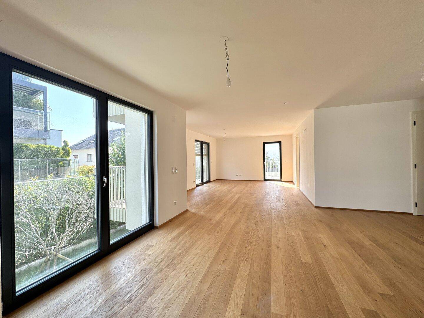 Lichtdurchfluteter Wohnbereich mit großen Fenstern, Holzboden und Zugang zu einem Außenbereich.