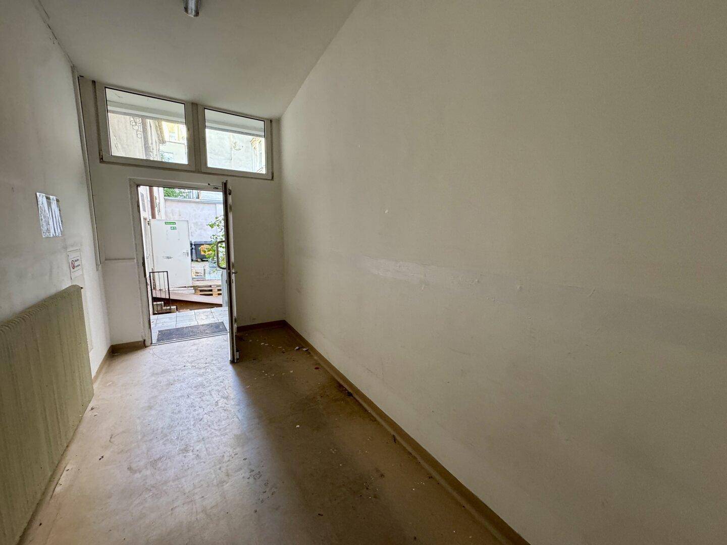 Eingangsbereich mit offener Tür nach draußen, kleinen Fenstern und einfachem Bodenbelag.