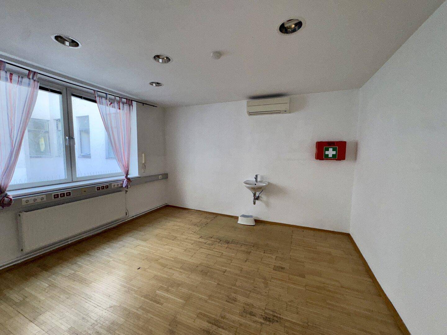 Heller Raum mit Holzboden, großem Fenster, Heizkörper, Waschbecken und Klimaanlage.
