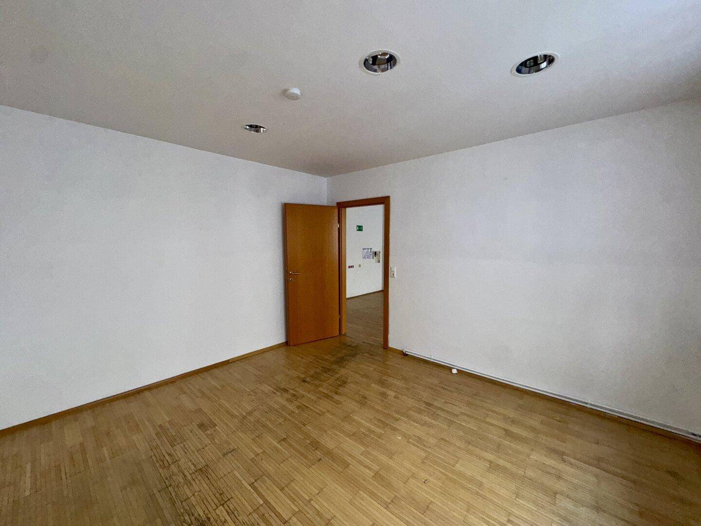 Leerer Raum mit Holzboden, weißen Wänden und offener Holztür, ausgestattet mit Einbaustrahlern.