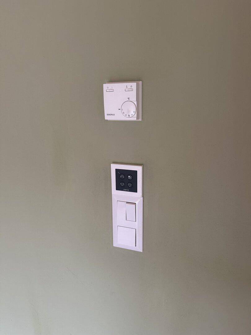 Wand mit einem Thermostat zur Temperaturregelung und einem modernen Lichtschalter.