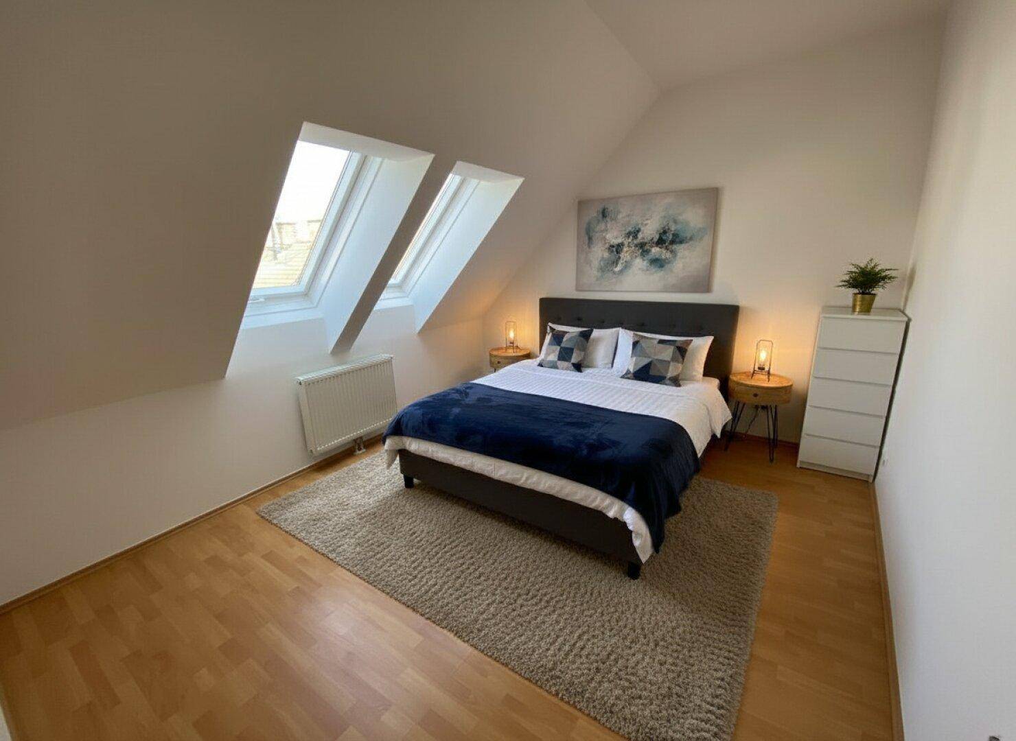 Gemütliches Schlafzimmer im Dachgeschoss mit schrägen Wänden, zwei Dachfenstern und modernem Doppelbett.