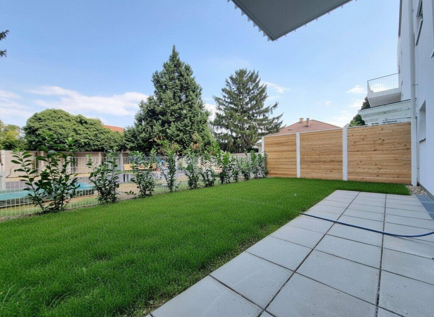 Gepflegter Gartenbereich mit Rasenfläche, Hecke und Holzzaun, umgeben von hohen Bäumen.
