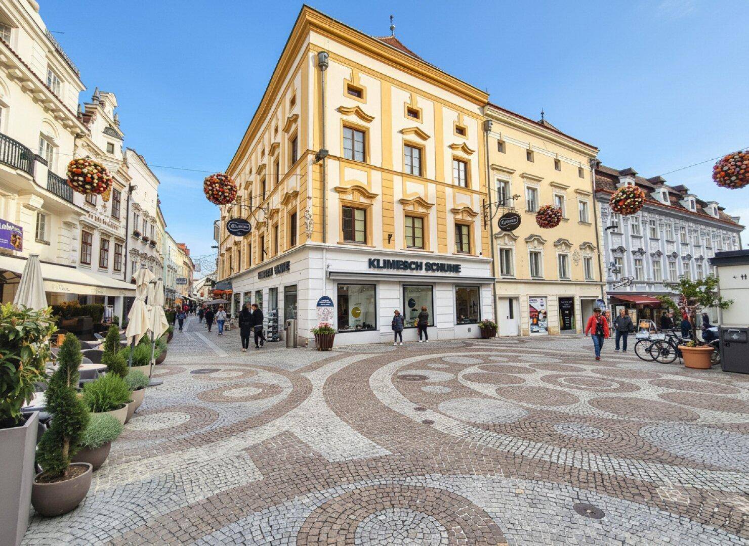 Belebte Einkaufsstraße mit historischen Gebäuden, gepflastertem Platz und festlicher Dekoration unter blauem Himmel.
