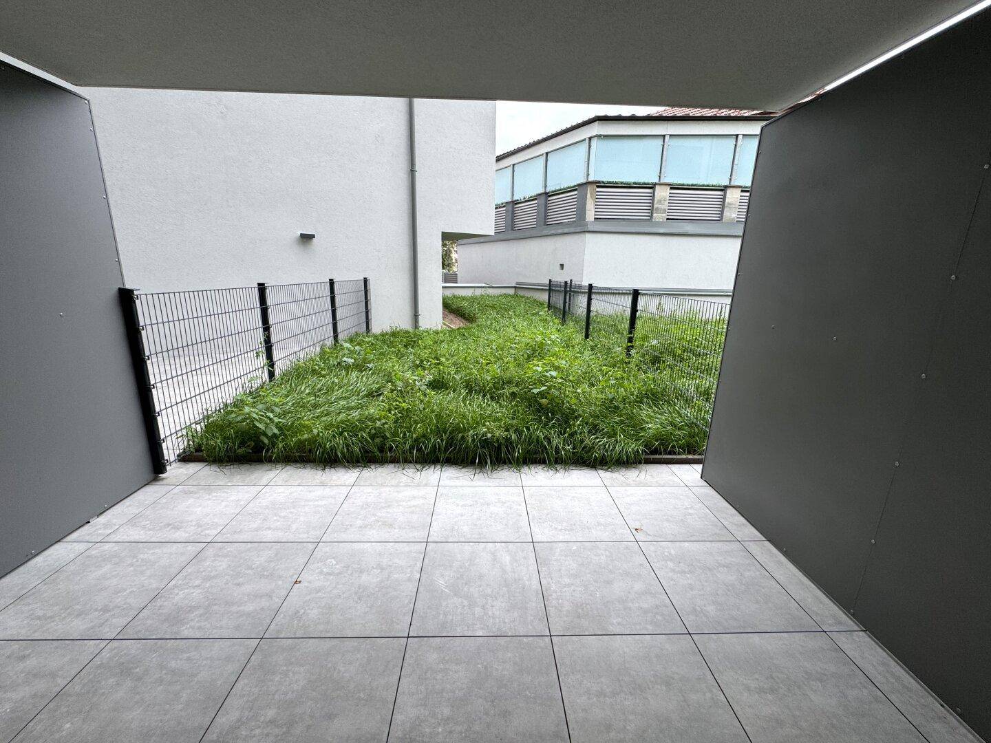 Eine geflieste Terrasse mit Blick auf eine kleine Grünfläche, umgeben von Zäunen.