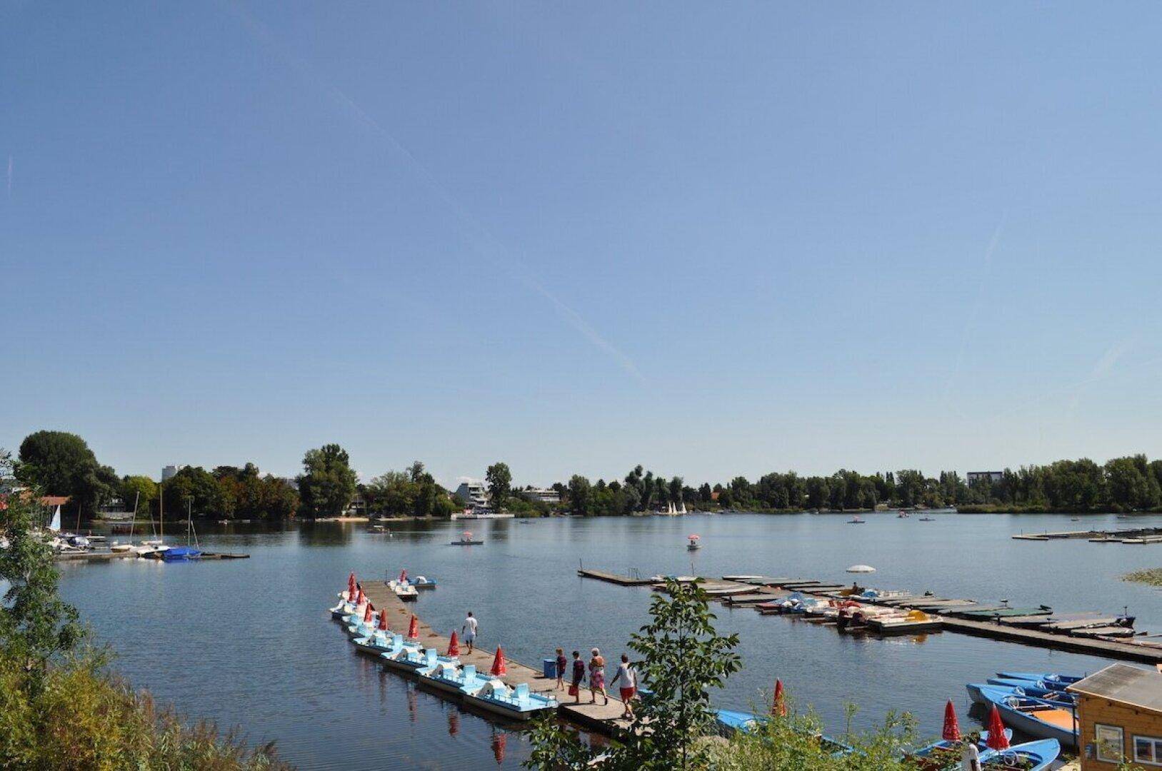 Malerischer Seeblick mit einem Steg, bunten Tretbooten und Menschen, die die sonnige Atmosphäre genießen.