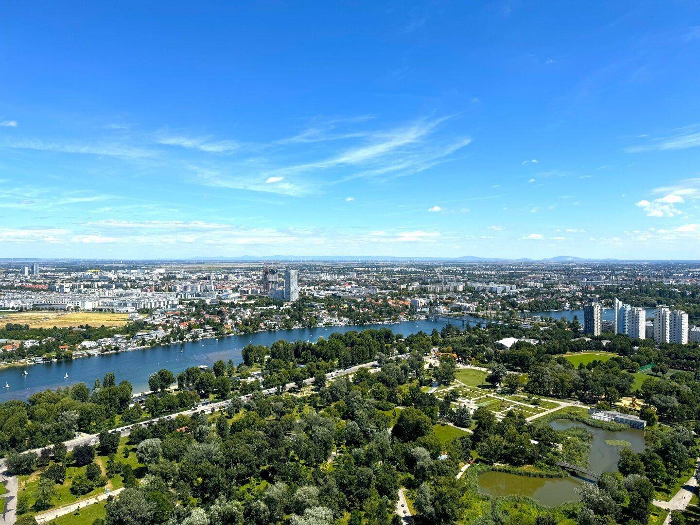 Weitläufige Luftaufnahme der Stadt mit Fluss, ausgedehnten Grünflächen und Wohngebäuden unter blauem Himmel.