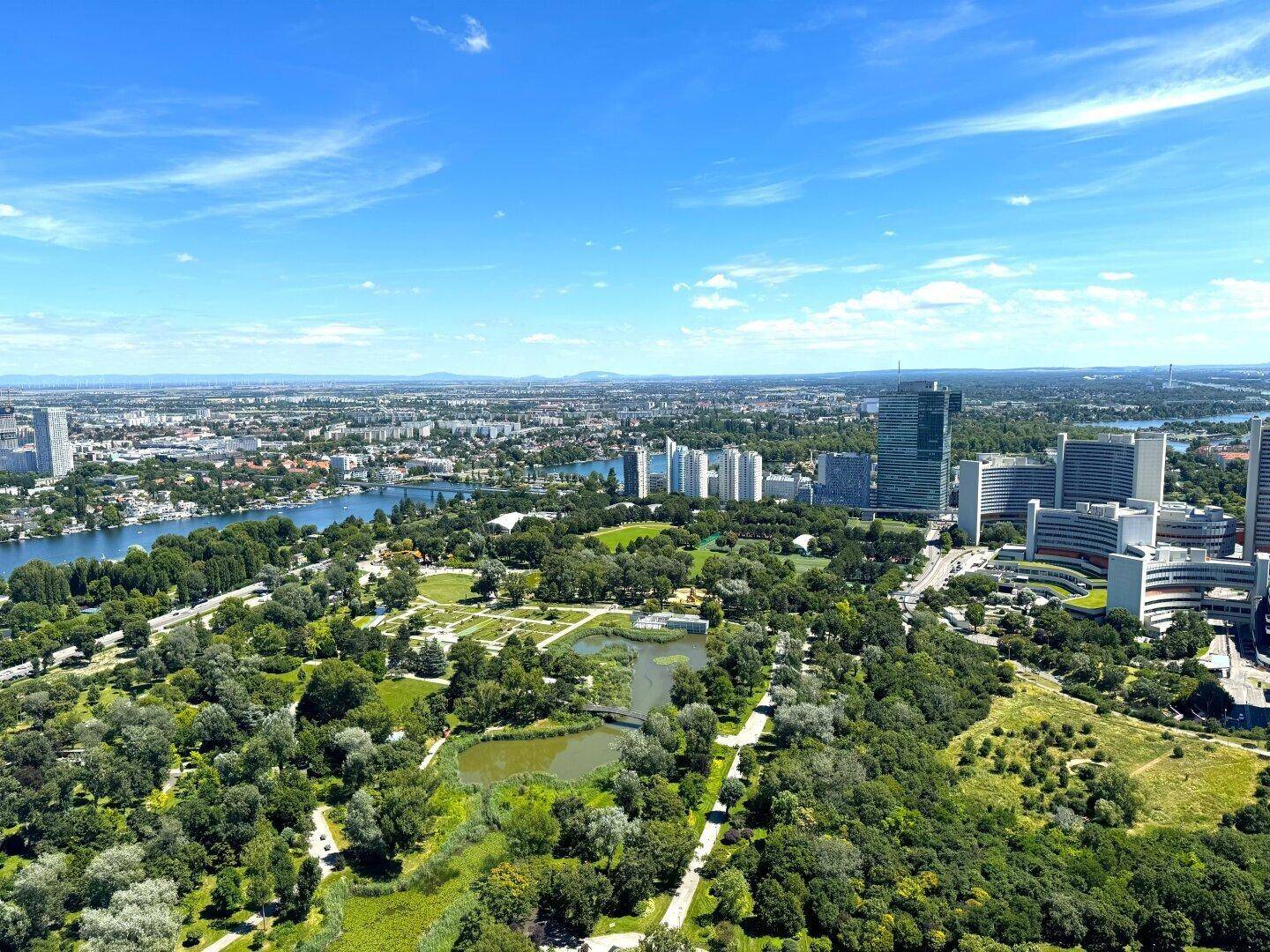 Weitläufige Luftaufnahme der Stadt mit Fluss, ausgedehnten Parkanlagen und modernen Gebäuden unter blauem Himmel.