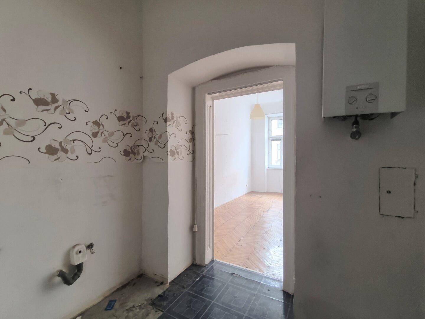 Küche im Rohzustand mit Gastherme und Zugang zu einem weiteren Zimmer.