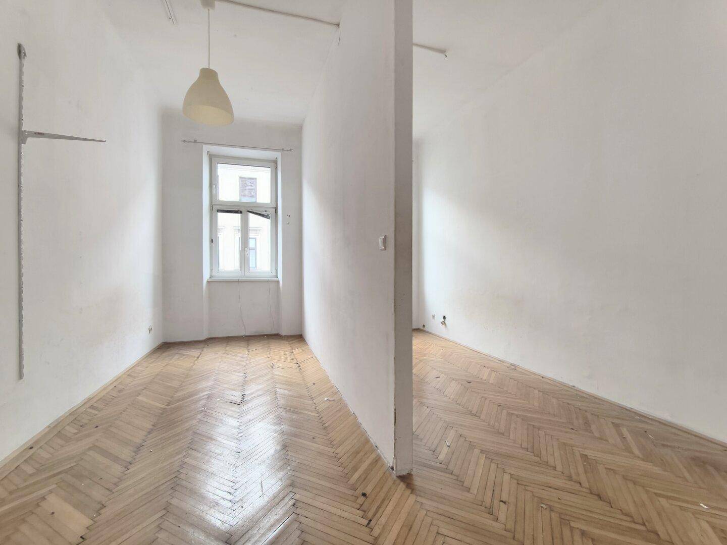 Leerer Raum mit Fischgrätparkett, Fenster und einer Trennwand, die den Bereich unterteilt.