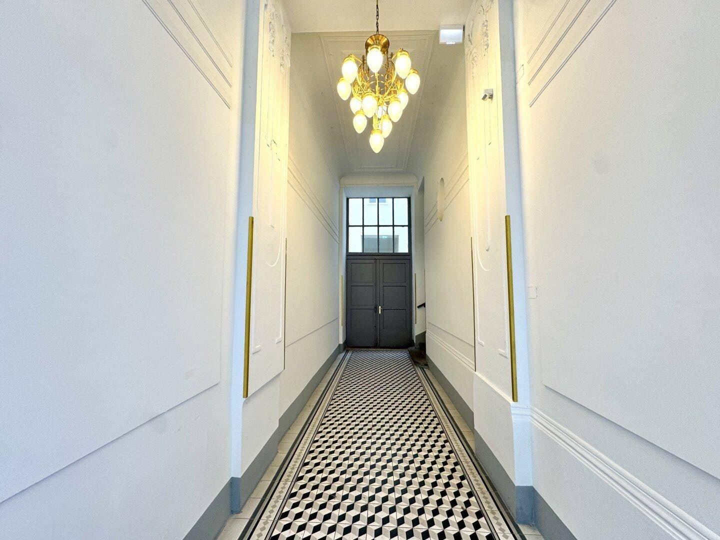 Langer, schmaler Flur mit auffälligem Schachbrettmuster-Boden und einem eleganten Kronleuchter.