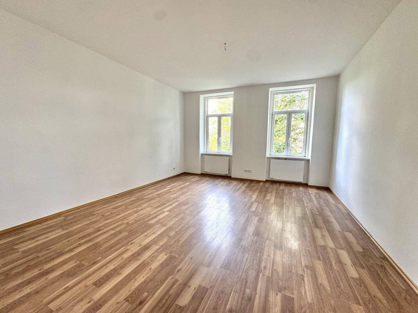 Heller, leerer Raum mit Holzboden und zwei großen Fenstern, die viel Tageslicht hereinlassen.