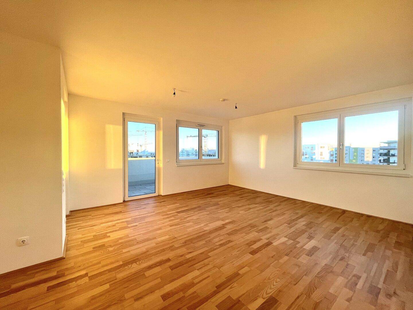 Helles, leeres Wohnzimmer mit Parkettboden und Zugang zu einem Balkon.