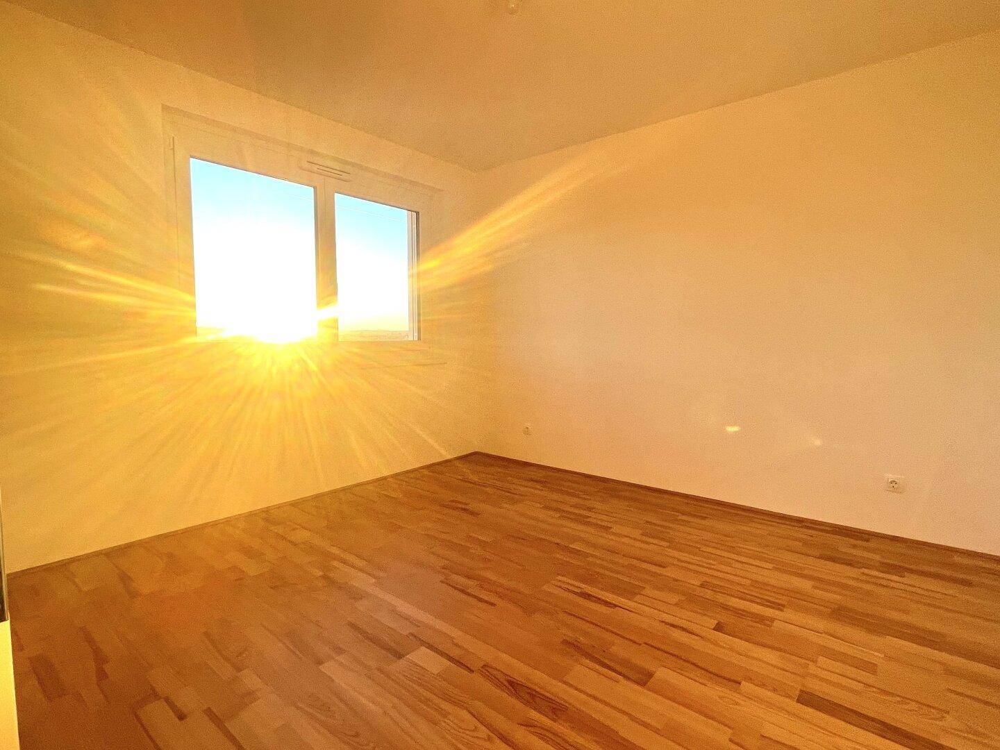 Helles Schlafzimmer mit Parkettboden und großem Fenster, das viel Sonnenlicht hereinlässt.