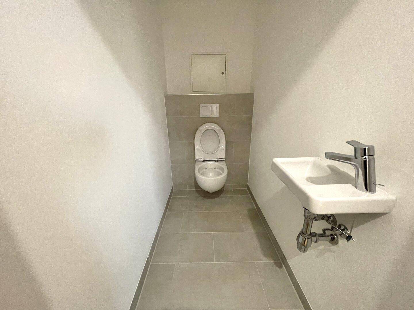 Kleines Gäste-WC mit wandhängender Toilette, kleinem Waschbecken und grauen Fliesen.