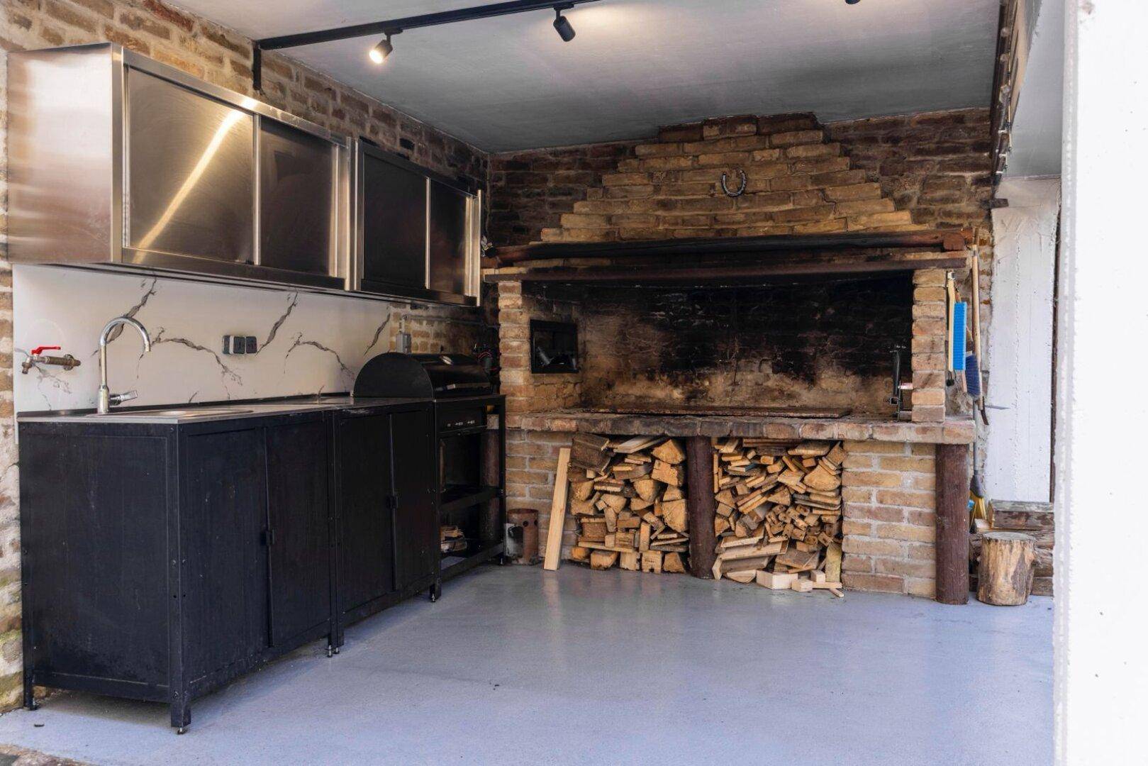 Gut ausgestattete Außenküche mit Edelstahlmöbeln, gemauertem Grill und Holzvorrat.