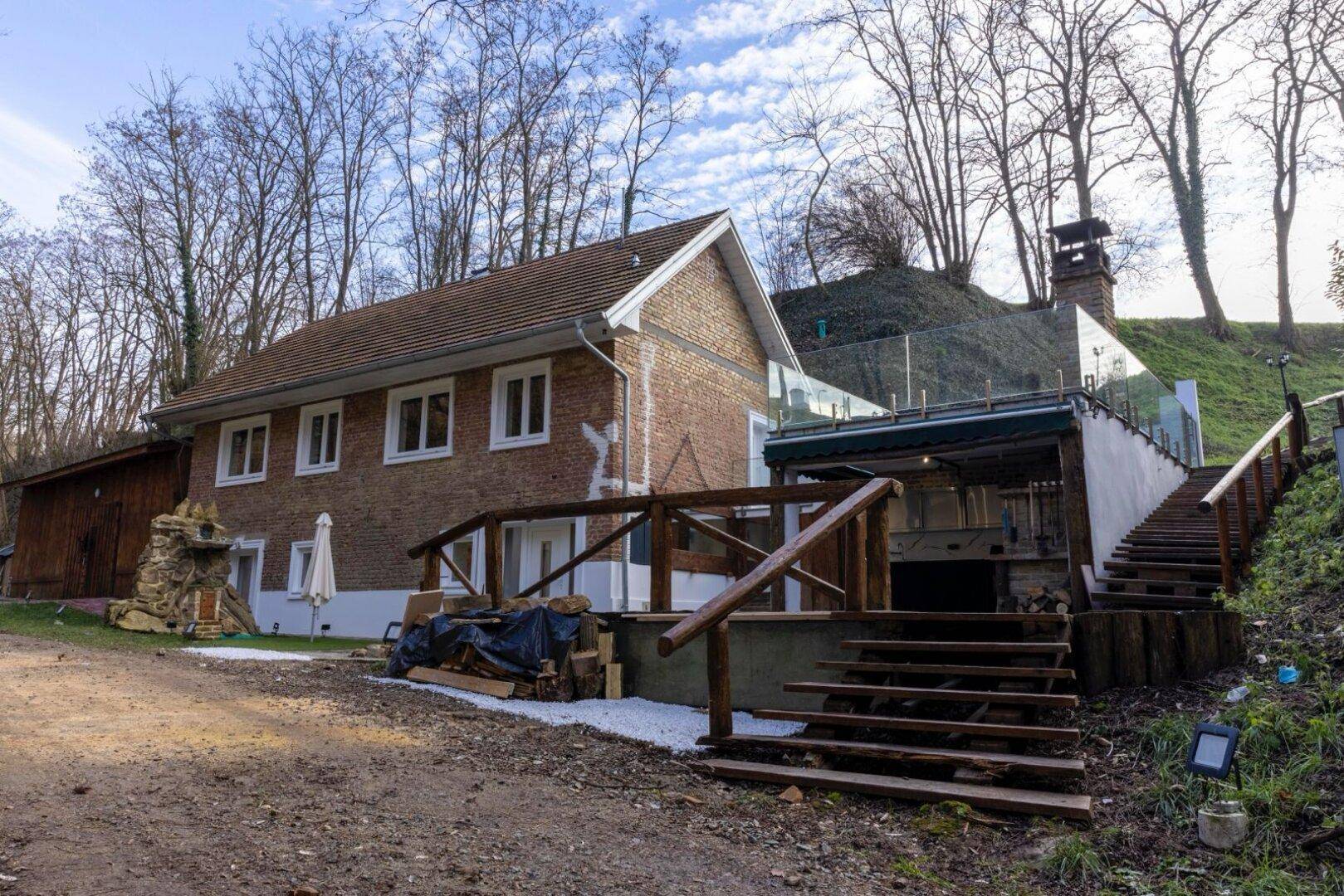 Seitenansicht des Ziegelhauses mit Holztreppen, einer überdachten Außenküche und Glasgeländer.