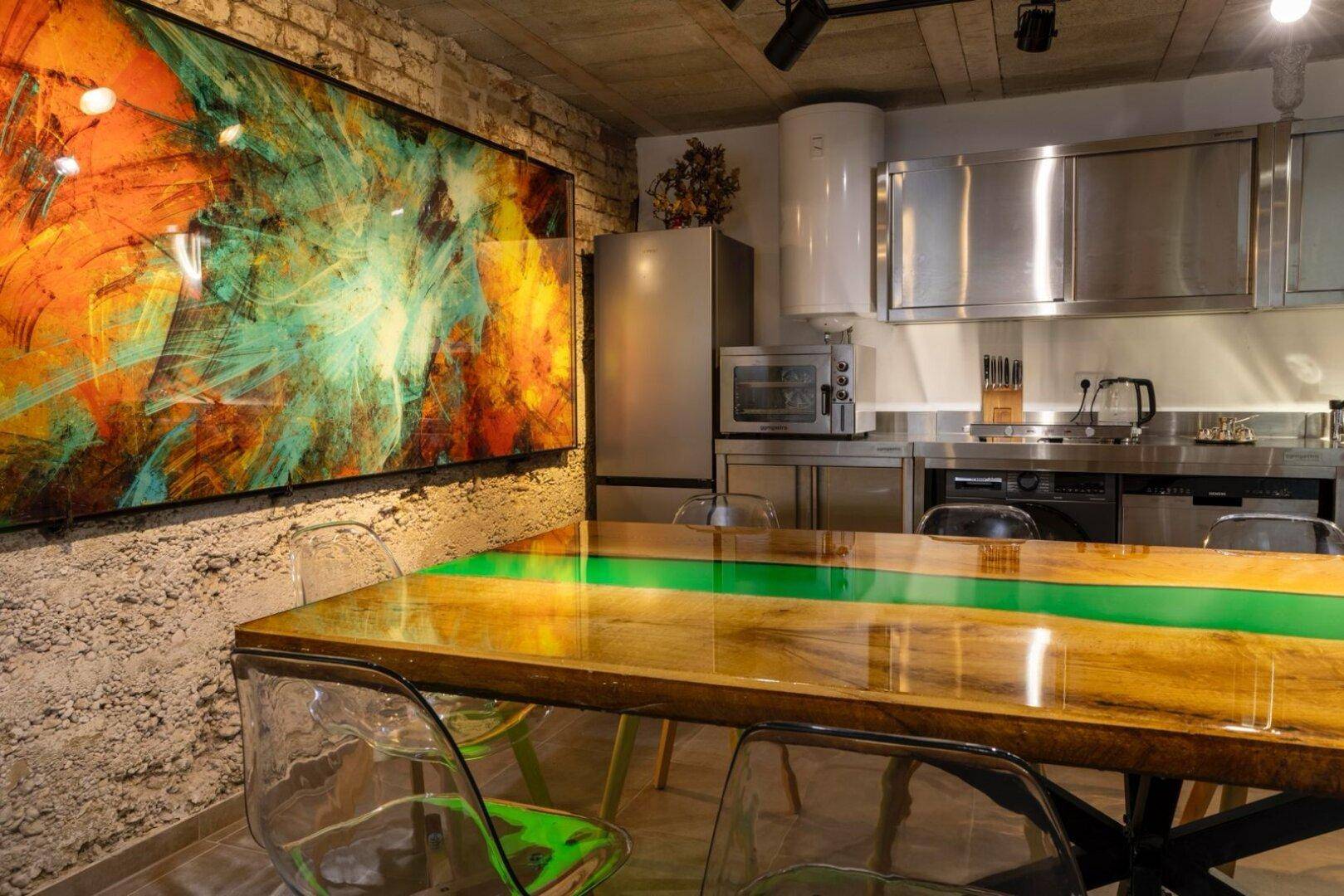 Moderne Edelstahlküche mit großem Esstisch und farbenfrohem Kunstwerk an der Wand.