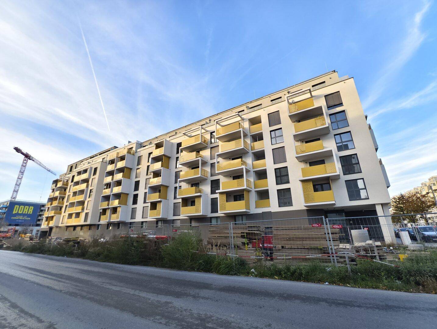 Modernes Mehrfamilienhaus im Bau mit vielen gelben Balkonen und einem Baukran im Hintergrund.
