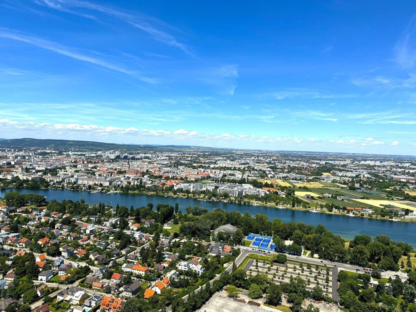 Luftaufnahme einer Stadt mit einem Fluss, Wohngebieten und weitläufigen Grünflächen unter blauem Himmel.