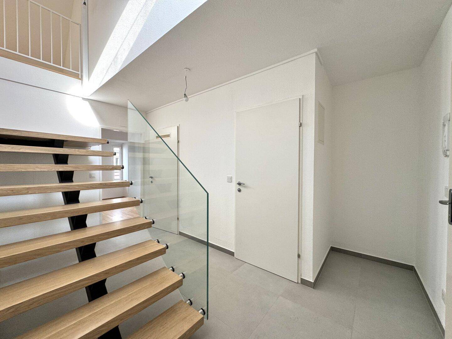 Moderner Eingangsbereich mit Holztreppe, Glasgeländer und Fliesenboden.