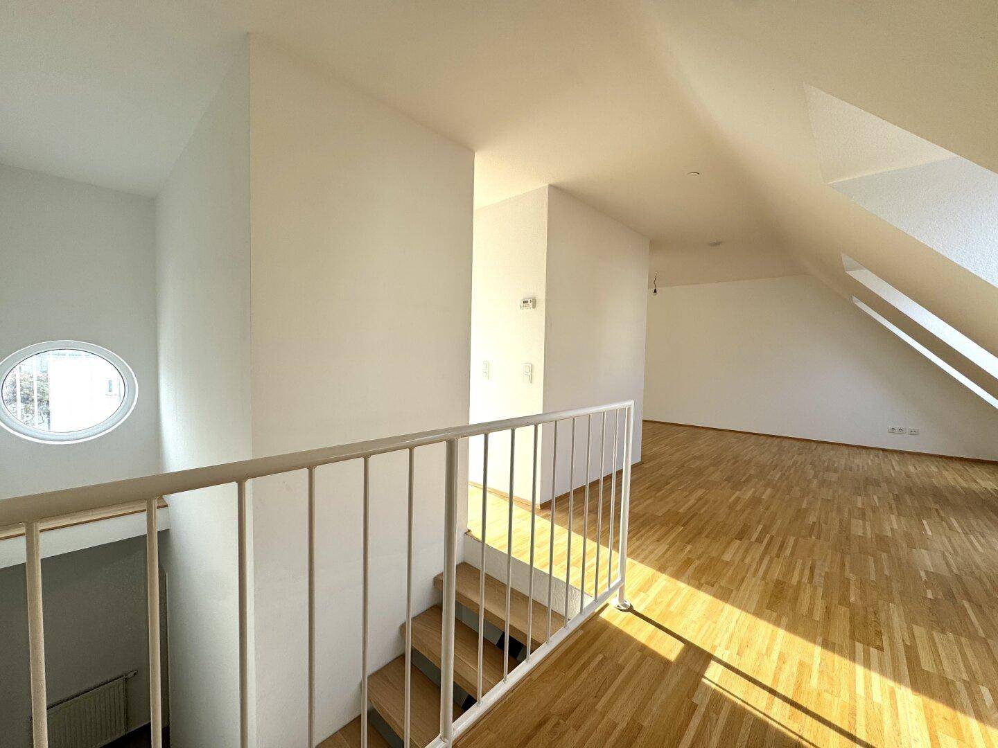 Heller Flurbereich mit Holztreppe, Geländer und runden Fenstern unter Dachschrägen.