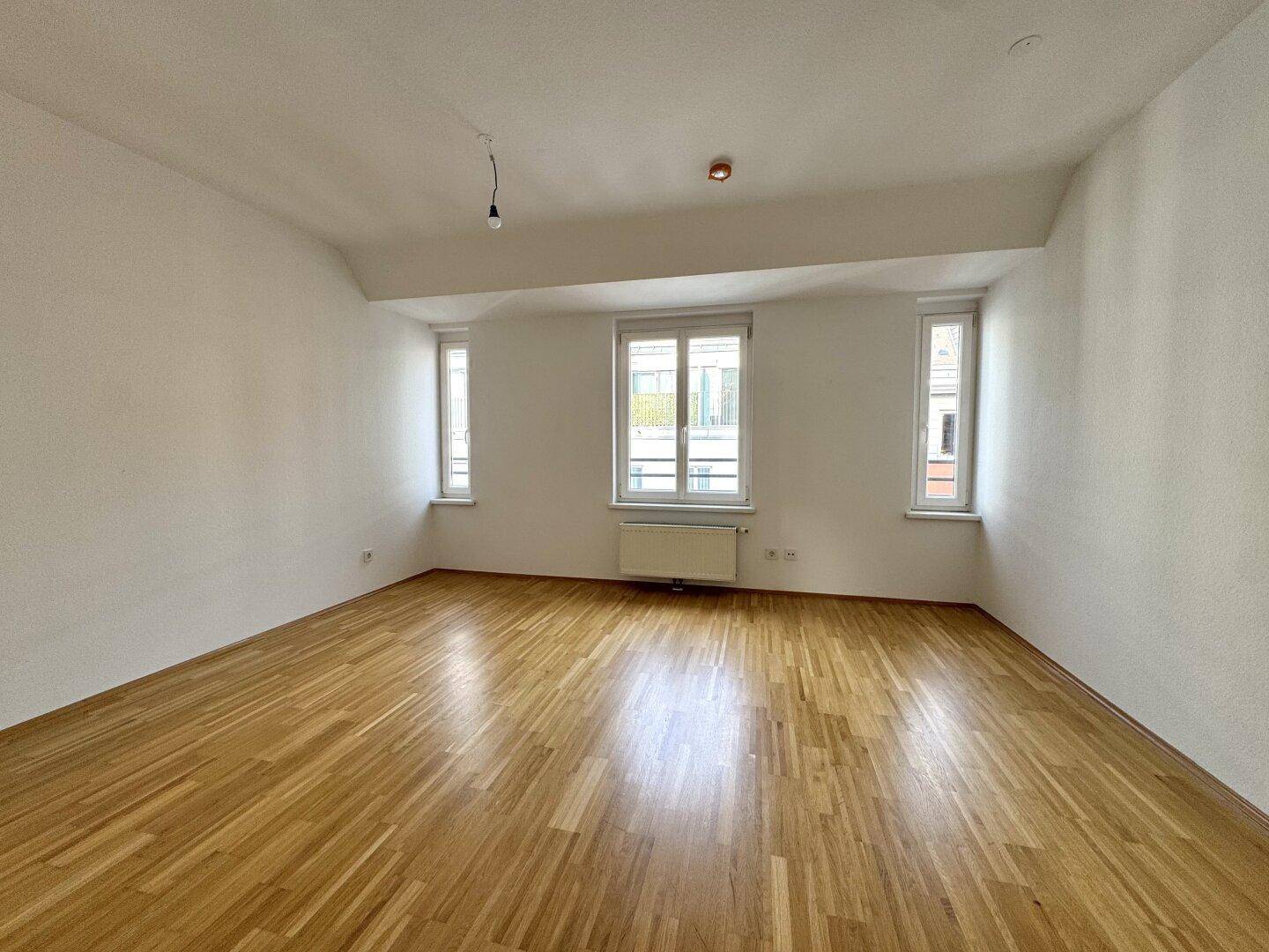 Geräumiges, leeres Schlafzimmer mit Holzboden und drei Fenstern, hell und freundlich.