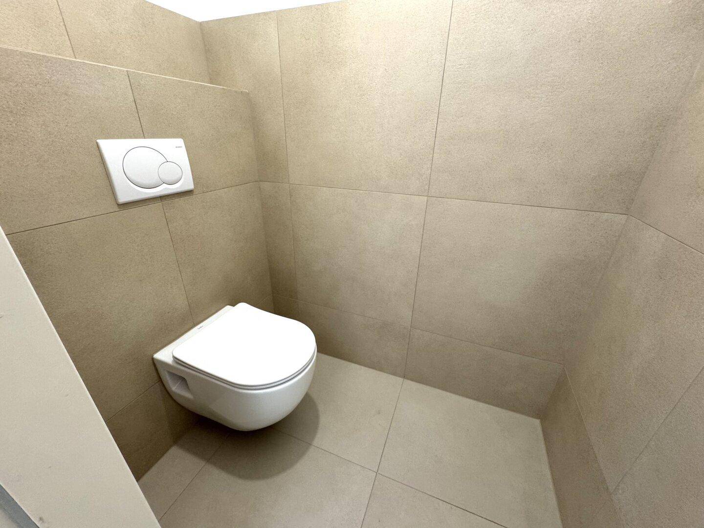 Separates WC mit wandhängender Toilette und beigefarbenen Fliesen.