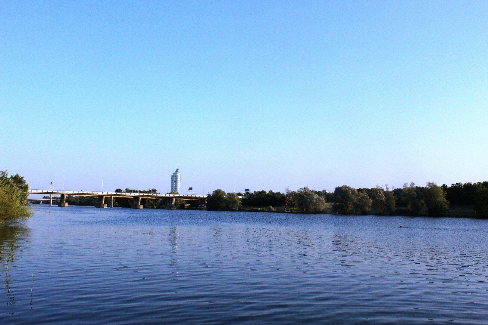 Ruhiger Flusslauf mit einer Brücke im Hintergrund und einem markanten Gebäude am Horizont.