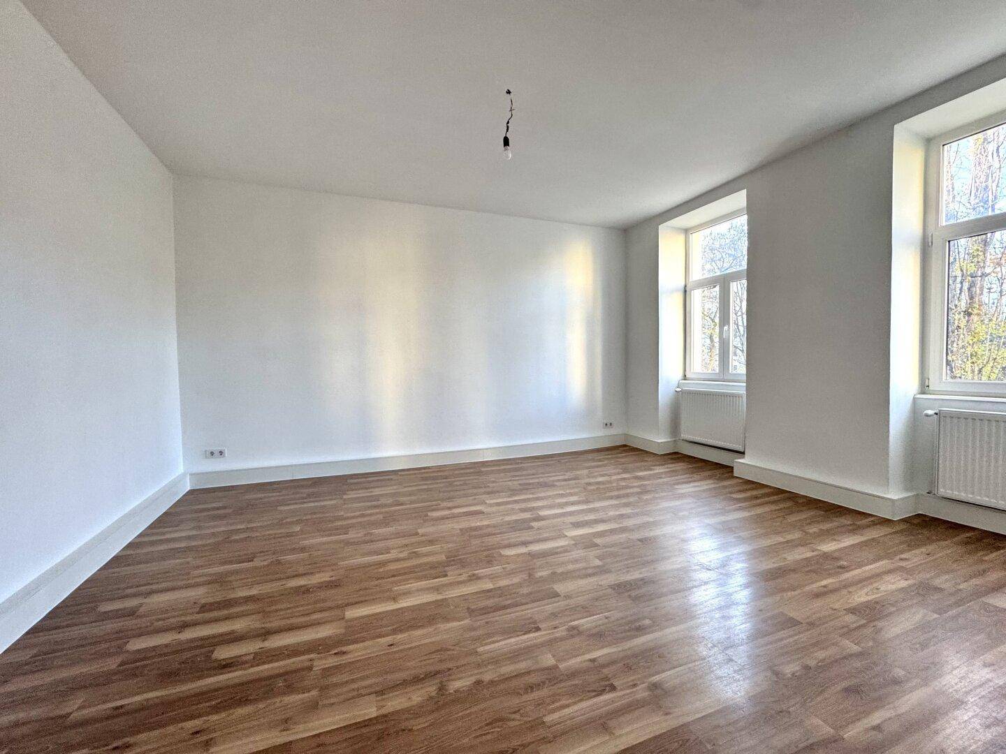 Leeres Wohnzimmer mit Holzboden und viel Tageslicht durch die hohen Fenster.
